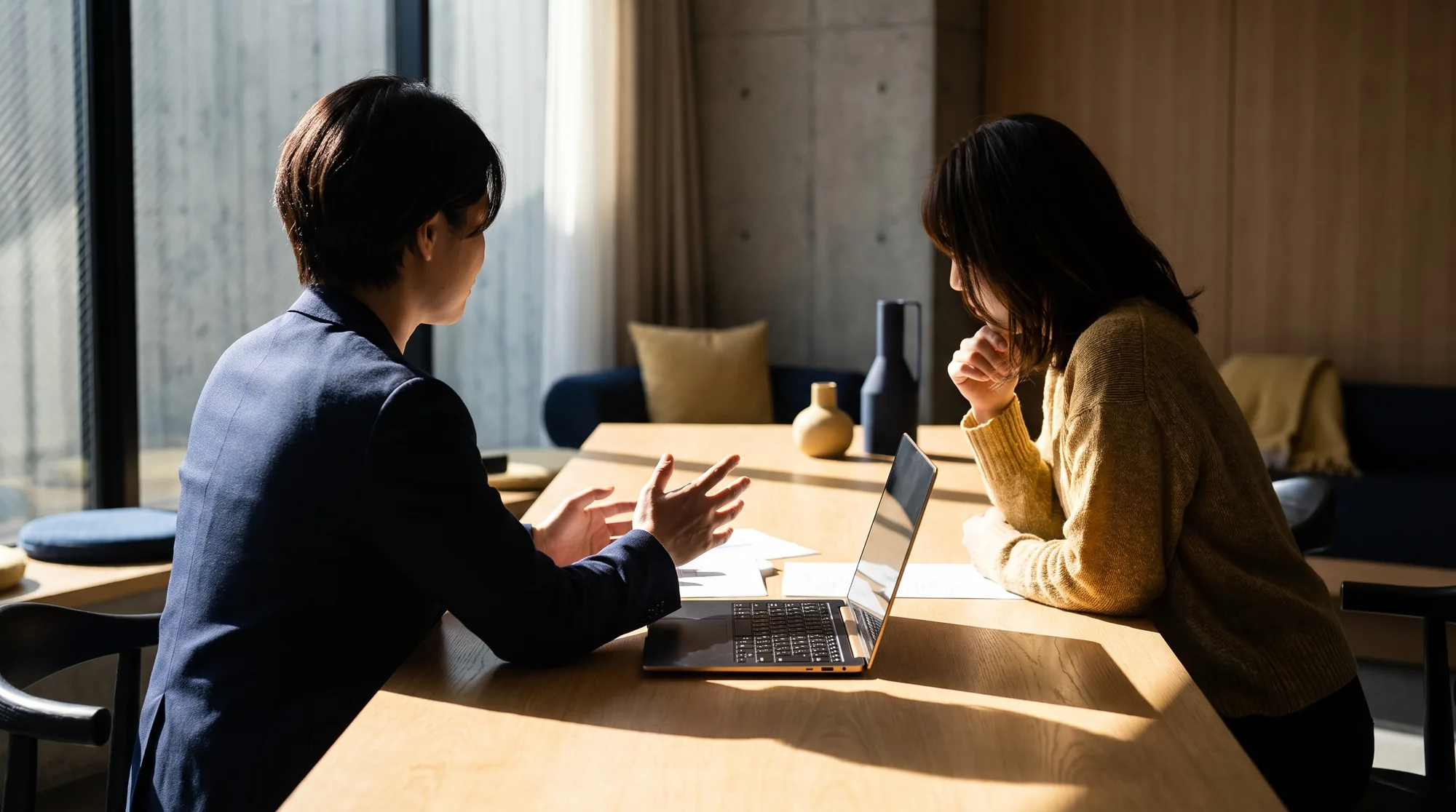 明るい相談室で、二人の専門家が資料とノートパソコンを前に向き合って話し合っている様子(後ろ姿)